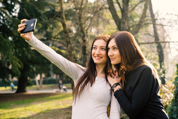 Portrait of young sisters having fun taking a selfie with smartphone outdoors in a park. - Fotó, kép
