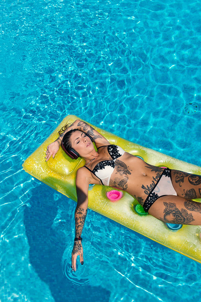 Sensual tattooed woman wearing bikini relaxing while listening to music with earphones inside swimming pool.  - Fotografie, Obrázek
