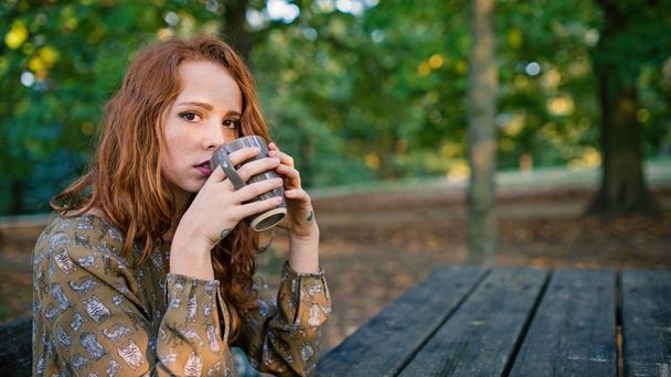 Intimate portrait of young redhead woman drinking from a cup outdoors in a park in autumn. Shallow depth of field. - Фото, зображення