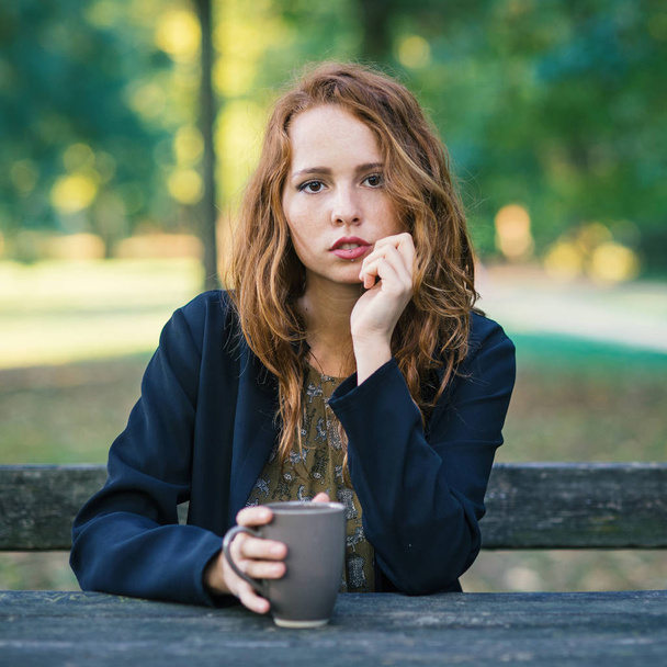 Intense portrait of young redhead woman drinking from a cup outdoors in a park in autumn. - Photo, image