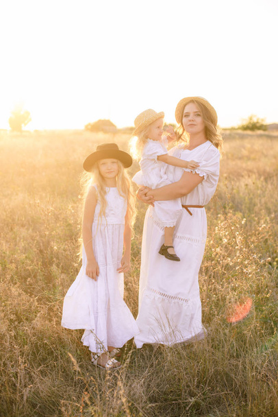 Young mother with daughters with blond hair in white dresses at sunset in the summer in a field of countryside - Foto, imagen
