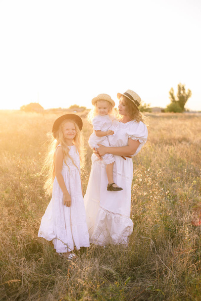 Young mother with daughters with blond hair in white dresses at sunset in the summer in a field of countryside - Фото, зображення