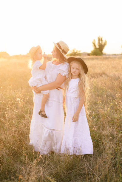 Young mother with daughters with blond hair in white dresses at sunset in the summer in a field of countryside - Foto, immagini