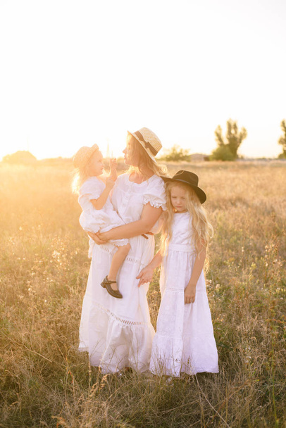 Madre joven con hijas con cabello rubio en vestidos blancos al atardecer en el verano en un campo de campo
 - Foto, Imagen