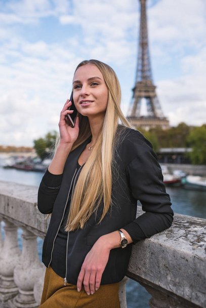 Młoda uśmiechnięta kobieta rozmawiająca przez telefon z wieżą Eiffla na tle, Paryż, Francja. - Zdjęcie, obraz