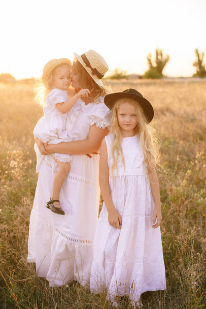 Young mother with daughters with blond hair in white dresses at sunset in the summer in a field of countryside - Foto, Bild