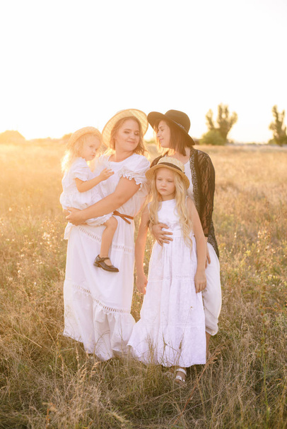 A young mother with her daughters and an aunt with blond hair in white dresses at sunset in the summer in a field of countryside. Several girls from the same family in the summer in the field - Fotoğraf, Görsel
