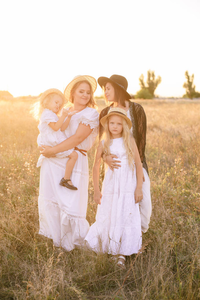 A young mother with her daughters and an aunt with blond hair in white dresses at sunset in the summer in a field of countryside. Several girls from the same family in the summer in the field - Fotoğraf, Görsel