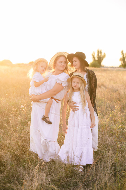 A young mother with her daughters and an aunt with blond hair in white dresses at sunset in the summer in a field of countryside. Several girls from the same family in the summer in the field - Foto, afbeelding