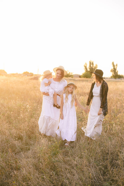 A young mother with her daughters and an aunt with blond hair in white dresses at sunset in the summer in a field of countryside. Several girls from the same family in the summer in the field - Zdjęcie, obraz