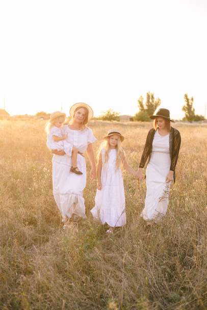 A young mother with her daughters and an aunt with blond hair in white dresses at sunset in the summer in a field of countryside. Several girls from the same family in the summer in the field - Fotografie, Obrázek