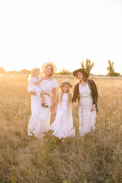 A young mother with her daughters and an aunt with blond hair in white dresses at sunset in the summer in a field of countryside. Several girls from the same family in the summer in the field - Photo, Image