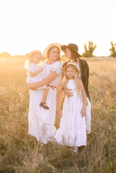 Una joven madre con sus hijas y una tía con el pelo rubio en vestidos blancos al atardecer en el verano en un campo. Varias chicas de la misma familia en el verano en el campo
 - Foto, Imagen