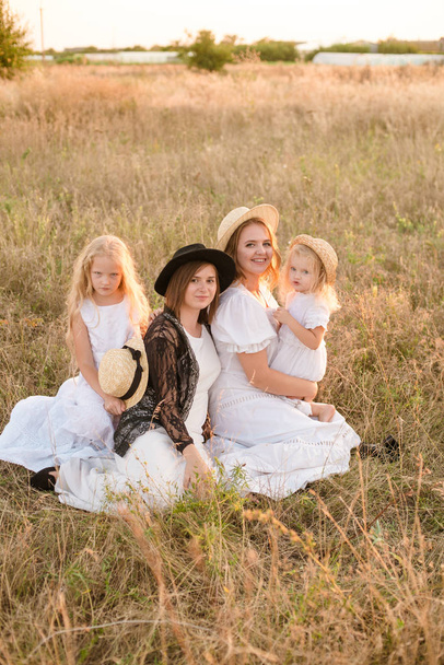 A young mother with her daughters and an aunt with blond hair in white dresses at sunset in the summer in a field of countryside. Several girls from the same family in the summer in the field - Photo, image