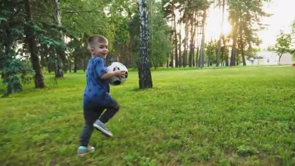 A cute little boy holding a soccer ball, running away over green grass and smiling happily. Slow motion shot - Footage, Video