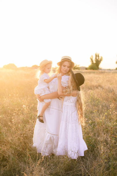 Young mother with daughters with blond hair in white dresses at sunset in the summer in a field of countryside - Photo, image