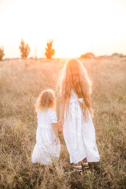 Cute girl with blond long hair with a younger sister in a summer field at sunset with a white dress - Foto, imagen