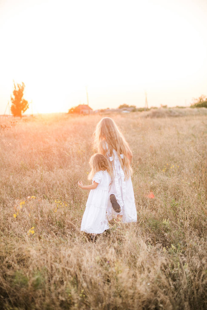 Χαριτωμένο κορίτσι με ξανθιά μακριά μαλλιά με μια μικρότερη αδελφή σε ένα καλοκαιρινό χωράφι στο ηλιοβασίλεμα με ένα λευκό φόρεμα - Φωτογραφία, εικόνα