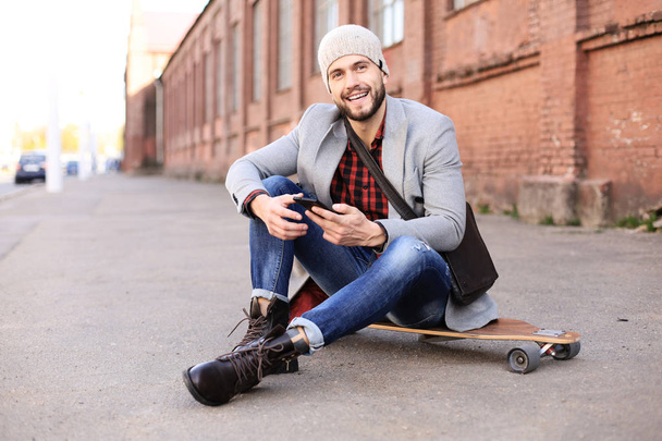 Handsome young man in grey coat and hat sitting on the longboard on the street in the city. Urban skateboarding concept. - Photo, Image