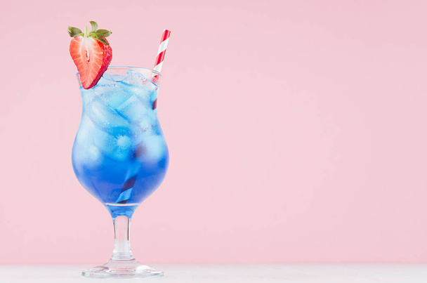 Summer blue alcohol drink with curacao liqueur, ice cubes, strawberry slice, straw in glamour glass on soft light pink background. - Fotografie, Obrázek