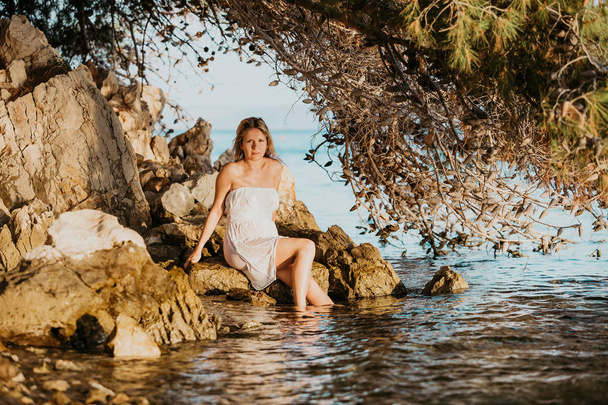 Hermosa joven embarazada con vestido mojado blanco relajarse en las rocas cerca del agua de mar Verano al atardecer Concepto Amor y embarazo
 - Foto, Imagen