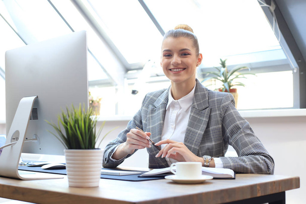 Portrait of a cheerful young businesswoman sitting at the table in office and looking at camera. - Foto, Imagem