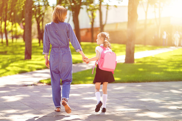 Parent and pupil of primary school go hand in hand. Woman and girl with backpack behind the back. Beginning of lessons. First day of fall - 写真・画像