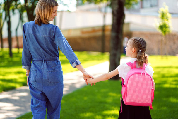 Parent and pupil of primary school go hand in hand. Woman and girl with backpack behind the back. Beginning of lessons. First day of fall - Фото, зображення