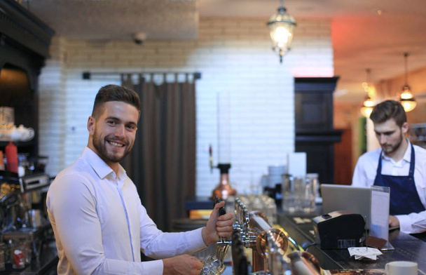 Guapo camarero está sonriendo y llenando un vaso de cerveza mientras está de pie en el mostrador del bar en el pub
. - Foto, imagen