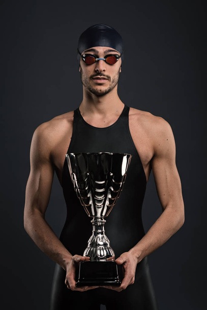 Professional athlete swimmer studio portrait against dark background holding  cup. Victory concept. - Foto, Imagen