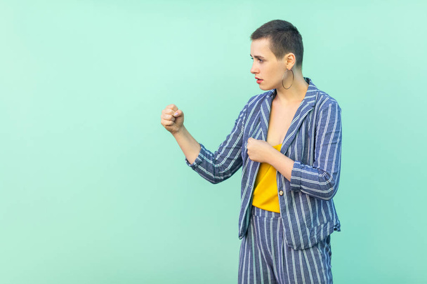 side view of serious beautiful short hair young stylish woman in casual striped suit standing with boxing fists and ready to attack  on light green background - 写真・画像