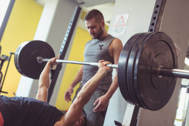 Young man lifting the barbell in the gym with friend helping him. Concept of active lifestyle. - Foto, Imagem