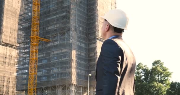 Portrait of a successful young handsome engineer, architect, builder, businessman, wearing a white helmet, in a suit, holding a notepad, skyscraper background and building. - Felvétel, videó