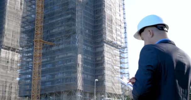 Portrait of a successful young handsome engineer, architect, builder, businessman, wearing a white helmet, in a suit, holding a notepad, skyscraper background and building. - Filmagem, Vídeo