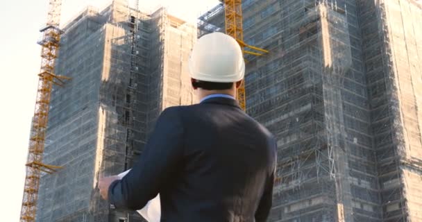 Portrait of a successful young handsome engineer, architect, builder, businessman, wearing a white helmet, in a suit, holding a notepad, skyscraper background and building. - Footage, Video