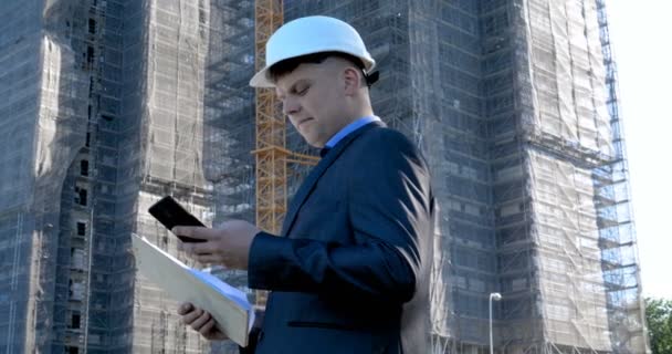 Retrato de um jovem e bem sucedido engenheiro bonito, arquiteto, construtor, empresário, vestindo um capacete branco, em um terno, segurando um bloco de notas, fundo do arranha-céu e construção. - Filmagem, Vídeo