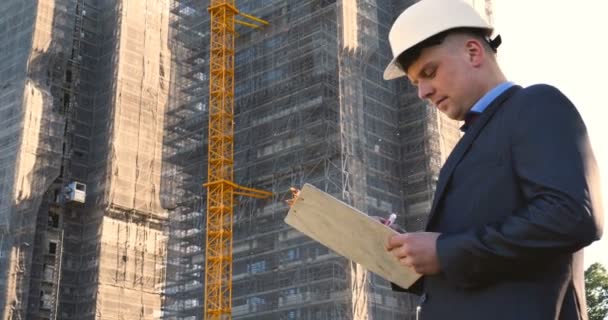 Portrait of a successful young handsome engineer, architect, builder, businessman, wearing a white helmet, in a suit, holding a notepad, skyscraper background and building. - Video