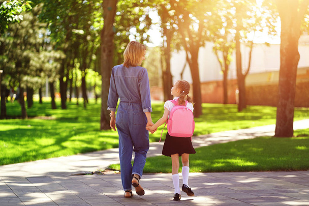 Parent and pupil of primary school go hand in hand. Woman and girl with backpack behind the back. Beginning of lessons. First day of fall - Foto, Imagen