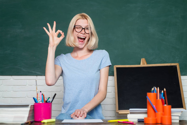 Portrait of funny young Caucasian female teacher wink and show OK. Young happy student. Portrait of Female University Student indoors. Beautiful teacher in classroom. - Φωτογραφία, εικόνα