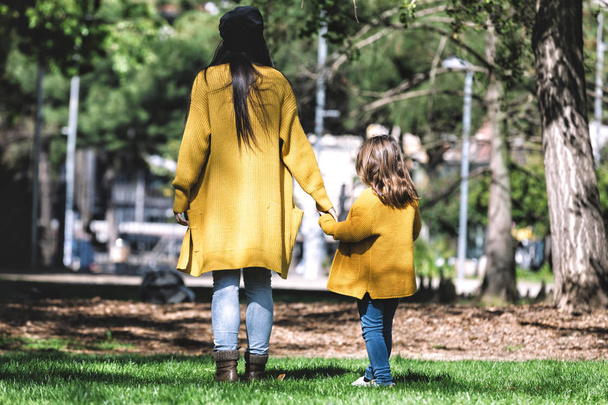 Rear view of mother and little daughter, dressed in yellow jacket, walking and holding hands in the park - Fotoğraf, Görsel