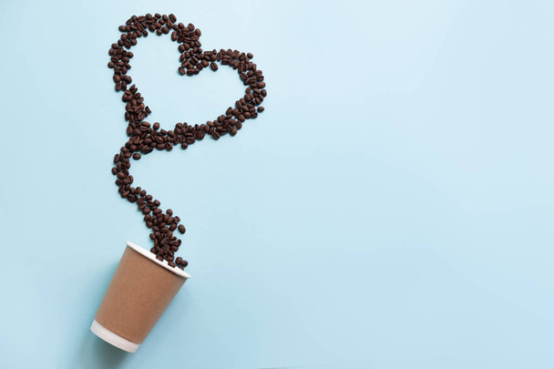 Κόκκους καφέ σε σχήμα καρδιάς και χάρτινο φλιτζάνι απομονώνεται σε μπλε φόντο - Φωτογραφία, εικόνα