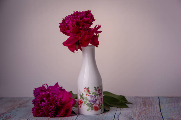 Una peonía roja está en un jarrón blanco con flores pintadas, dos peonías se encuentran una al lado de la otra sobre un fondo de madera
. - Foto, Imagen