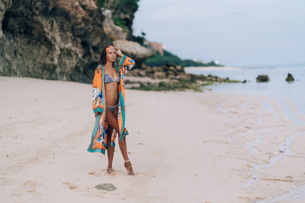 Beautiful sexy girl in swimwear walking on sandy beach with rock on background - Foto, Imagen