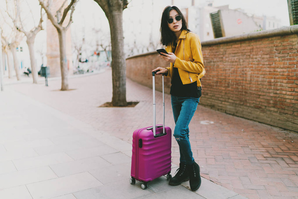 Travel blogger using internet on a mobile phone to find direction to destination. Asian hipster girl with a luggage using mobile application to call for a taxi while standing on a street. - 写真・画像