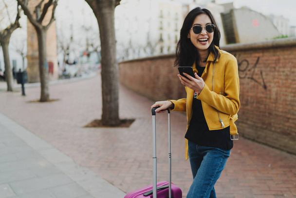 Young smiling woman traveler enjoying her first minutes in a new city and sharing the emotions with a friends online by a smartphone. Asian hipster girl reading messages on a mobile phone. - Fotó, kép