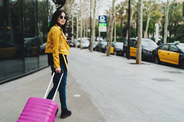 Young asian entrepreneur female with a suitcase answering business e-mail on the go to a taxi to the airport during important business trip. Traveler girl buying tickets online by a mobile phone. - 写真・画像