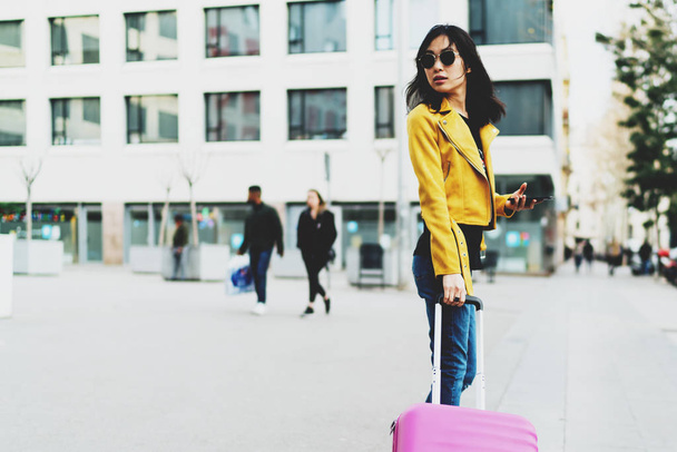 Side view photo of model look asian hipster female wearing sunglasses and modern outfit looking back while standing with a pink suitcase and a mobile phone in her hand on a blurred street background. - Фото, зображення