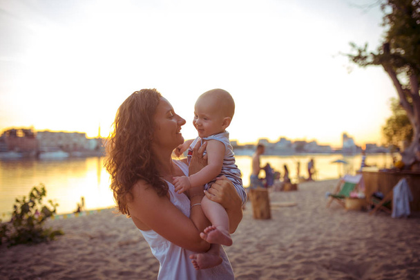 Νεαρή λευκή γυναίκα, όμορφη μητέρα κρατά ένα παιδί στην αγκαλιά της, η μαμά αγκαλιάζει το γιο της, ενός έτους, στέκεται σε μια αμμώδη παραλία σε ένα πάρκο, υπαίθριο καφέ, παραλιακό καφέ το καλοκαίρι στο ηλιοβασίλεμα - Φωτογραφία, εικόνα