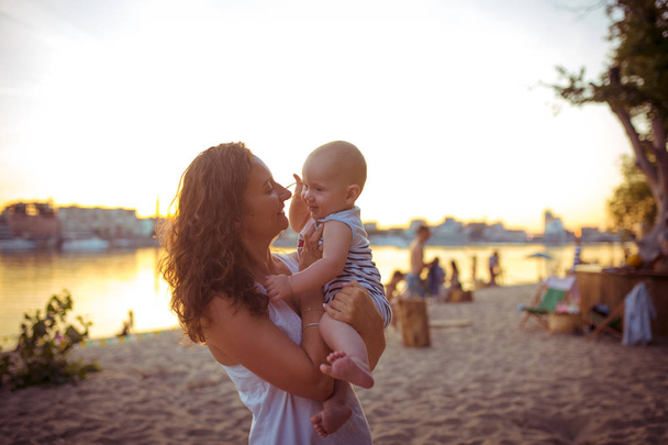 Young Caucasian woman, beautiful mother holds a child in her arms, mom hugs her one-year-old son standing on a sandy beach in a park, outdoor cafe, beach cafe in the summer at sunset - Photo, Image