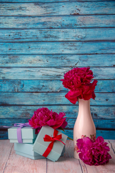 Yksi punainen pioni seisoo valkoisessa maljakossa, jossa on maalattuja kukkia, maljakon vieressä on kolme pientä lahjapakkausta, jotka on koristeltu jousilla ja kahdella pionilla, takana on sininen puinen tausta.
. - Valokuva, kuva